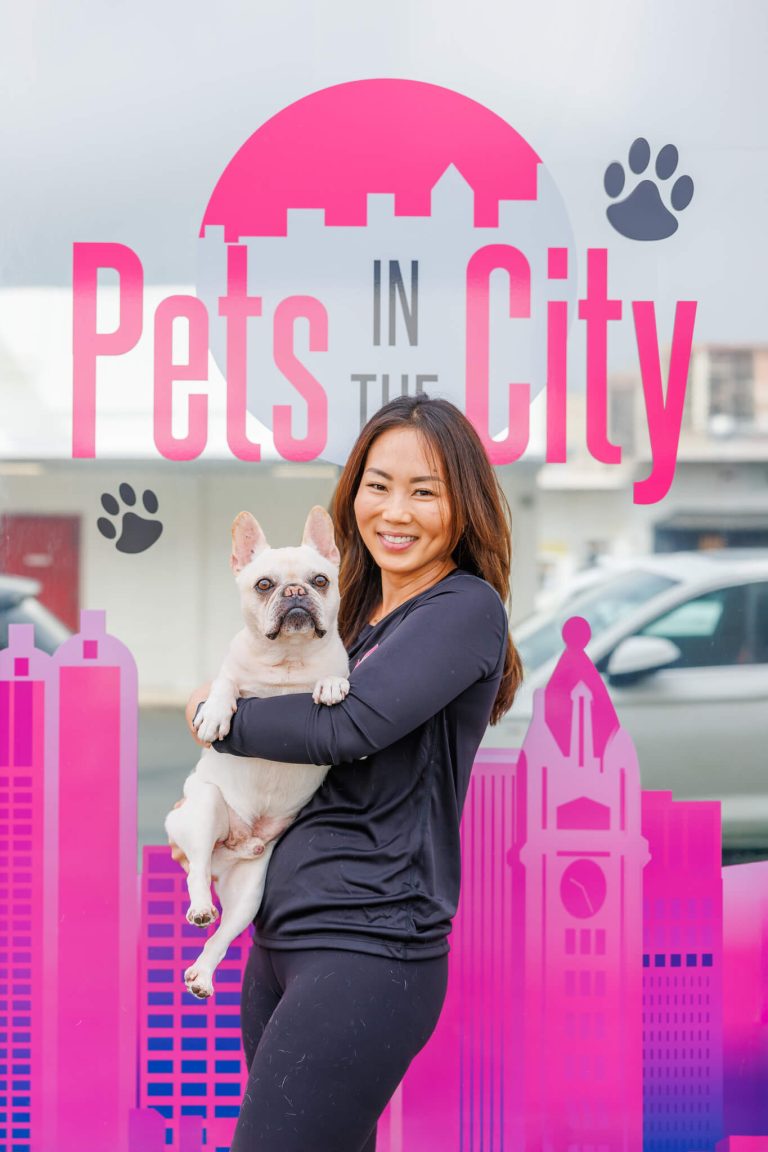 Ashley, owner of Pets in the City, holding Diesel, her French Bulldog, outside the facility with the Pets in the City logo in the background.